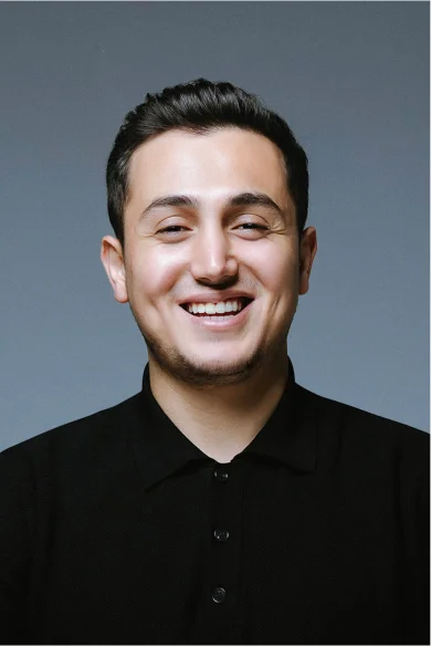 Cihat Ersin Aydın has short hair, a smiling face, and his hair is slightly styled upwards and straight. He is wearing a black T-shirt and smiling at the camera.